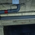 Canalisation métallique suspendue fixée au plafond d’un sous-sol en béton, utilisée pour l’évacuation des eaux usées dans un bâtiment, avec raccords visibles et isolation partielle