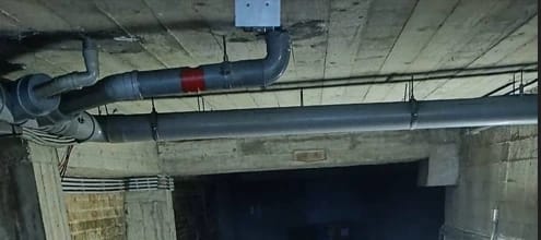 Canalisation métallique suspendue fixée au plafond d’un sous-sol en béton, utilisée pour l’évacuation des eaux usées dans un bâtiment, avec raccords visibles et isolation partielle